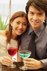 Couple sitting side by side, holding drinks and looking at camera