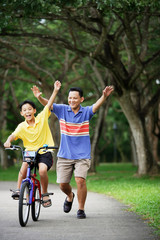 Boy on bicycle, father behind him, both with arms outstretched