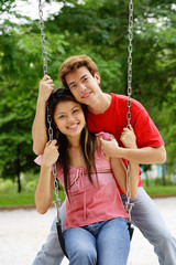 Couple in playground, woman sitting on swing, man behind her, smiling at camera