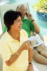 Woman holding coffee mug, man on lounge chair, using mobile phone