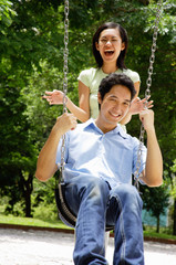 Man sitting on swing, woman standing behind him, laughing