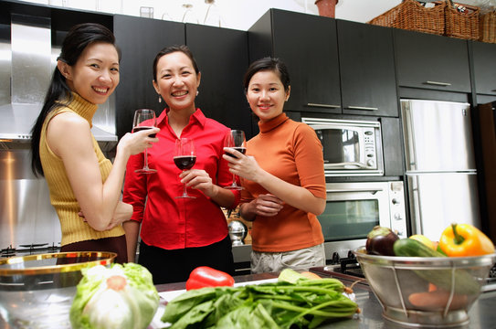 Three women in kitchen, holding wine glasses, looking at camera
