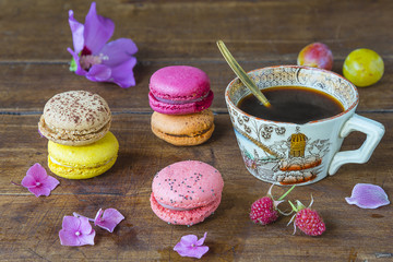 Multicolored french macaroons, cup of black coffee, several purple flowers and berries on old wooden background