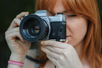 Young girl with orange hair taking pictures with an old camera retro style in a city park. She is happy to be able to use a reel camera again. 