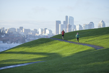 An adult man and woman running the paved path through Gasworks Park with the Seattle cityscape in the background. Washington State.