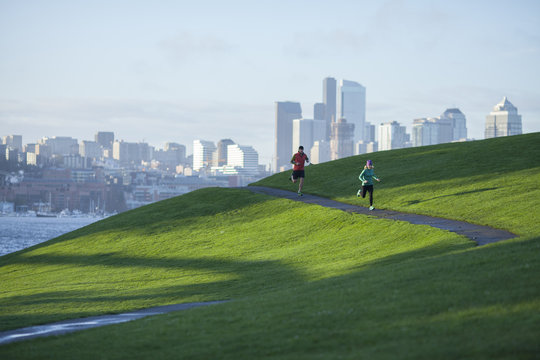 An adult man and woman running the paved path through Gasworks Park with the Seattle cityscape in the background. Washington State.