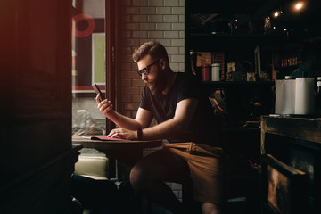 A bearded male is checking e-mail on a smartphone while waiting for the cup of tea in a cute city coffee shop with loft interior. Hipster guy in sunglasses is surfing the web on a mobile phone.