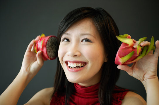 Woman holding dragon fruit halves next to her ears, smiling