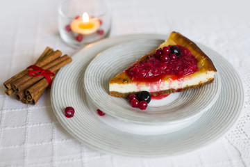 Чизкейк/творожный пирог с красными ягодами/красной смородиной на белой тарелке, палочки корицы, связанные красной нитью и горящая свеча и конфеты в красный обертках  - obrazy, fototapety, plakaty