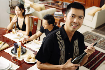 Man holding wine bottle, looking at camera, people in the background, sitting around dining table