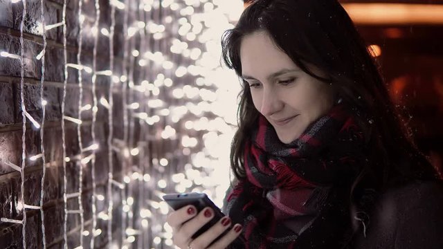 young attractive woman using smartphone in the falling snow at Christmas night standing near lights wall,