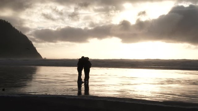 Unrecognizable silhouetted couple taking photo together with two birds flying by at the Pacific Ocean in Oregon during sunset.