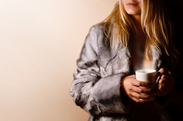 Stylish smiling woman in warm dress with a cup of tea or coffee on the background of beige wall....