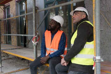 Two engineers wearing protective work wear talking sitting on scaffolding