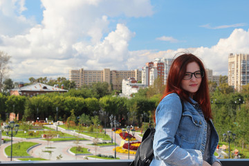 Fototapeta na wymiar Girl in glasses and a jean jacket on a background of city appearance and sky with clouds on a sunny day