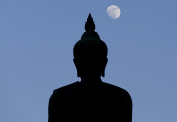 A moon shines in a sky over a large silhouetted Buddha statue in Bangkok, Thailand.