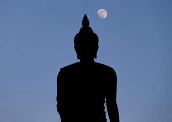 A moon shines in a sky over a large silhouetted Buddha statue in Bangkok, Thailand.
