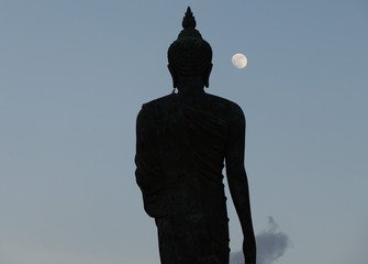 A moon shines in a sky over a large silhouetted Buddha statue in Bangkok, Thailand.