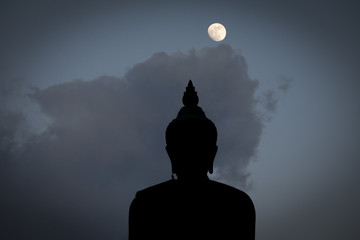 A moon shines in a sky over a large silhouetted Buddha statue in Bangkok, Thailand.