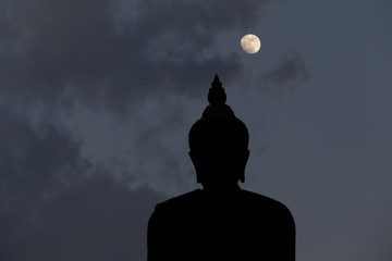 A moon shines in a sky over a large silhouetted Buddha statue in Bangkok, Thailand.