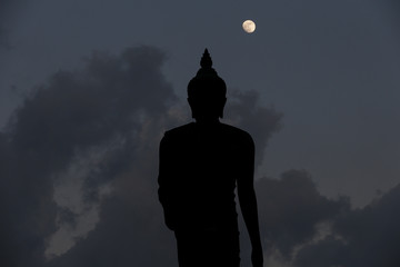 A moon shines in a sky over a large silhouetted Buddha statue in Bangkok, Thailand.