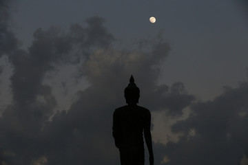 A moon shines in a sky over a large silhouetted Buddha statue in Bangkok, Thailand.