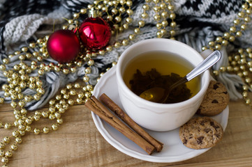 Fresh green tea with cinnamon sticks and homemade chocolate cookies, red christmas balls and golden beads, knitted blanket on wooden table