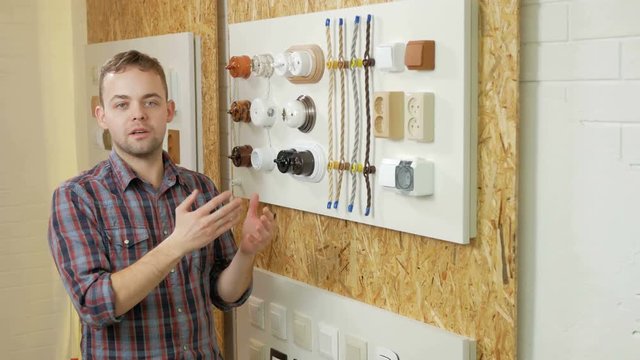 A young man tells and shows the switch sockets and wires of different colors and shapes. Samples are presented on a special stand. Looking at the camera