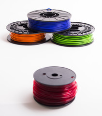 Stacked orange, blue and green 3D printer filament spools behind a blurry translucent red 3D printing filament spool