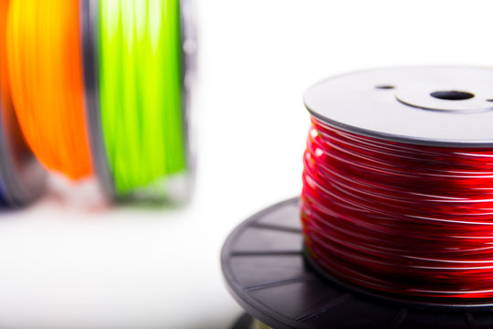 Partial view of translucent red 3d printing spool with blurry stacked blue and orange 3D printing filament spools in background