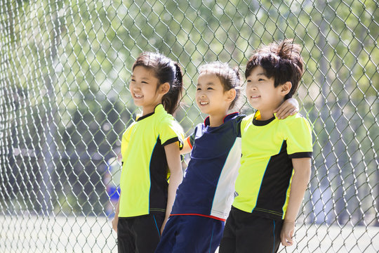 Happy children in sportswear leaning against chainlink fence