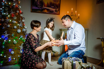 Mom and father give a small child Christmas gift on the tree background.