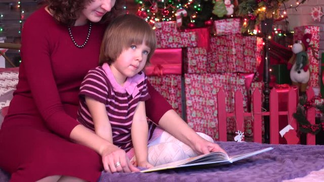 Attractive mother shows pictures in book to her cute little daughter over xmas background