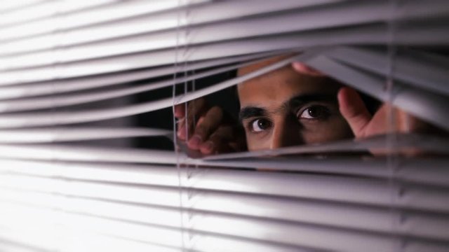 CU Man opening blinds and looking out at camera, side view