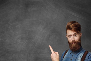 Hipster bearded man has idea, pointing his finger up, looking with amused expression, standing isolated in bottom right-hand corner of blank chalkboard with copy space for your promotional content