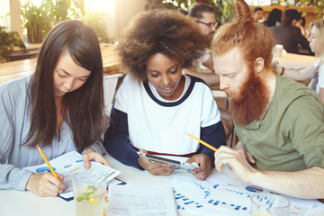 Asian girl filling in papers with graphs and diagrams while African woman sharing ideas with redhead bearded colleague on touch pad. Young businesspeople working on start-up project using tablet