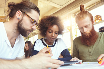 International group of three managers working together on new project, analyzing concept and plans, using digital tablet. Man in glasses explaining his vision pointing at touch pad screen with pencil