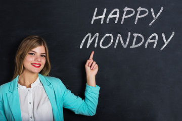 Beautiful young female student is pointing, looking at camera and smiling, standing against blackboard with text "happy monday"