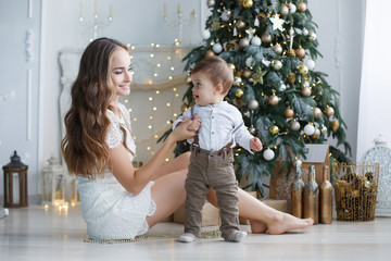 Beautiful brunette woman with long hair,spends the New Year holiday, sitting on a white floor with green Christmas tree along with baby son Christmas portrait of mother and child