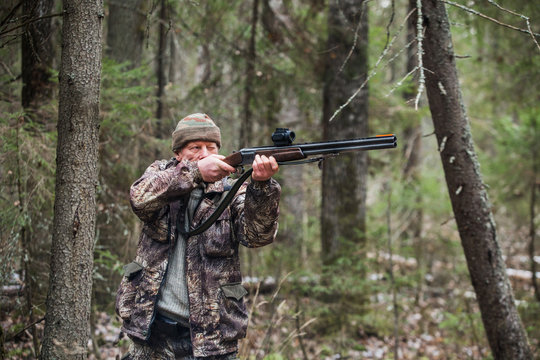 Male hunter shoots a gun. Hunter wearing a camouflage jacket and cap. A man of 50 years. On the ground lies a little of snow. Cloudy weather, autumn.