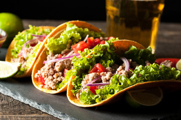 Photo of Mexican tacos with ground meat, beef, beans, onions and salsa on wooden background. Ketchup sauce and lime. A glass o beer in the background.