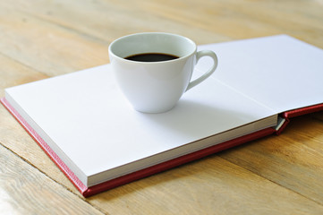 White Cup with coffee standing on the open book in red cover, wooden background.