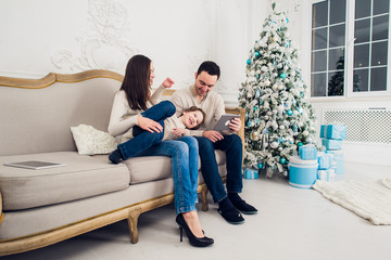 Obraz premium Cheerful family sitting in the living room having fun with the digital tablet that Santa Claus brought her, behind the decorated christmas tree