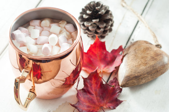 Hot chocolate, topped with marshmallow and served in a shiny copper mug. On a white wooden table with autumn leaves, pine cone and wooden table decoration.