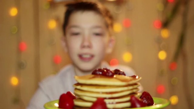 American pancakes. The boy is eating pancake with strawberry jam. On the background of colored lights.