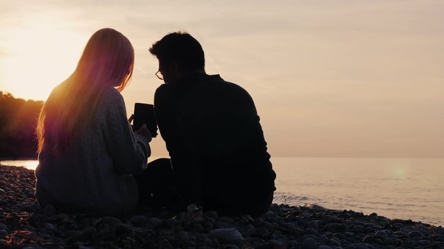 Silhouettes of man and woman sitting on pebble beach enjoying tablet. At sunset, the sea or a lake. Rear view, multiethnic couple