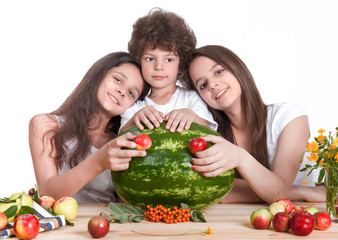 Three happy children are playing with a huge watermelon and fruit, sit at the table. The sisters put their heads on the shoulders of his brother. White background. Close-up.