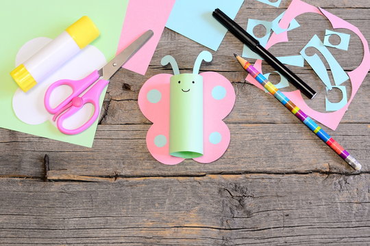 Funny paper butterfly, scissors, marker, glue stick, colored paper sheets and scraps, pencil on old wooden table. Children workplace. Summer children art craft project in kindergarten, camp or home