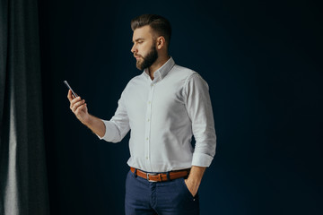View in profile of a young bearded businessman, dressed in a white shirt, standing in the center of the frame near the window on a dark-blue walls and looking at the screen of smartphone in his hand.