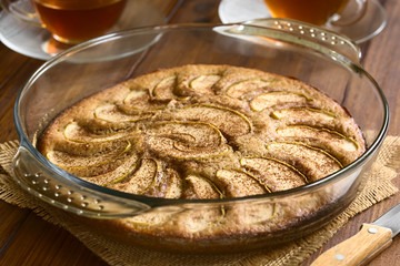 Homemade apple and cinnamon cake in glass dish with two cups of tea in the back, photographed with natural light (Selective Focus, Focus on the first apple slice in the middle of the cake)